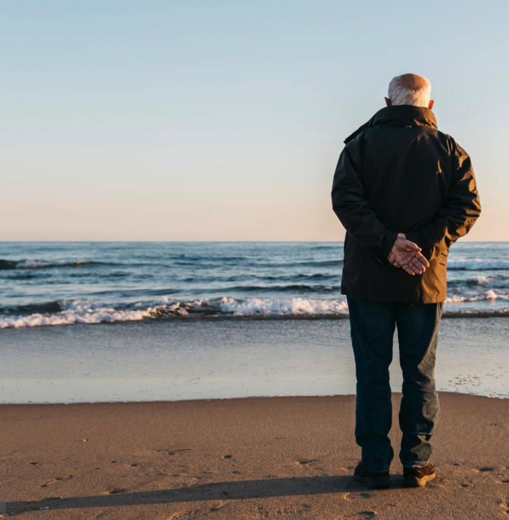 An older man standing alone on a beach, looking out at the ocean at sunset